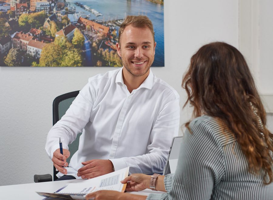 Das Bild zeigt einen freundlich Lächelnden Mitarbeiter der Personalabteilung, wie er ein Gespräch mit einer Mitarbeiterin führt. Es herrscht eine angenehme und offene Arbeitsatmosphäre.