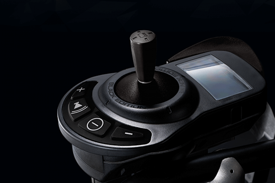 The image is black and white and shows a detail of the MAX-E wheelchair add-on drive. The focus is on the control unit. On the control unit you can see the ON/OFF button, the horn and the speed setting. The control unit is operated via a joystick. The display provides information about the current charge status. 