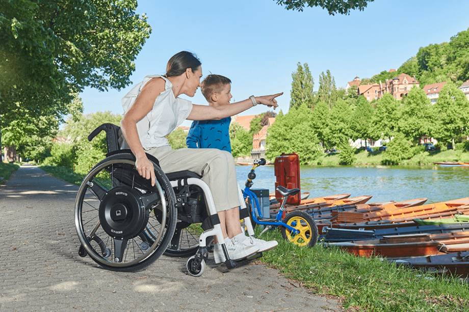Auf dem Bild ist eine Frau mittleren Alters zu sehen, die in einem weißen Rollstuhl mit schwarzen Radnabenantrieb zur Restkraftverstärkung am Ufer eines Flusses sitzt und einem Kind etwas zeigt.