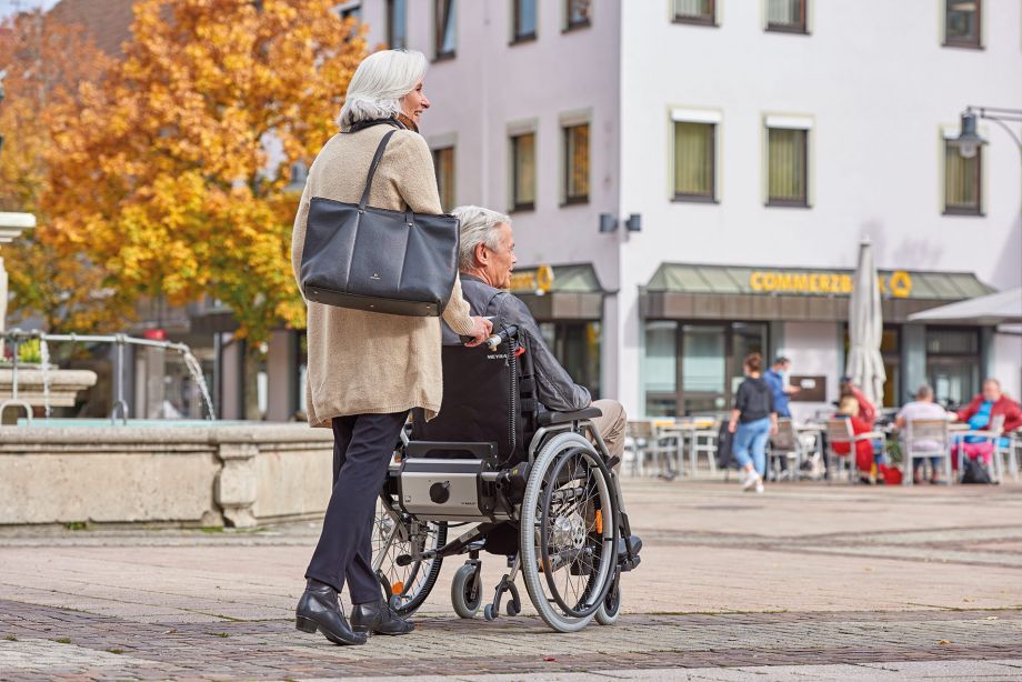 Das Bild zeigt eine ältere Seniorin mit grauen Haaren und großer Handtasche, wie sie einen älteren Senior mit ebenfalls grauen Haaren an einem Herbsttag durch die Stadt schiebt. An seinem manuellen Rollstuhl befindet sich unterhalb der Rückenlehne eine Brems- und Schiebehilfe V-MAX2 der Firma AAT Alber Antriebstechnik GmbH, welche sie beim Schieben und Bremsen unterstützt. 