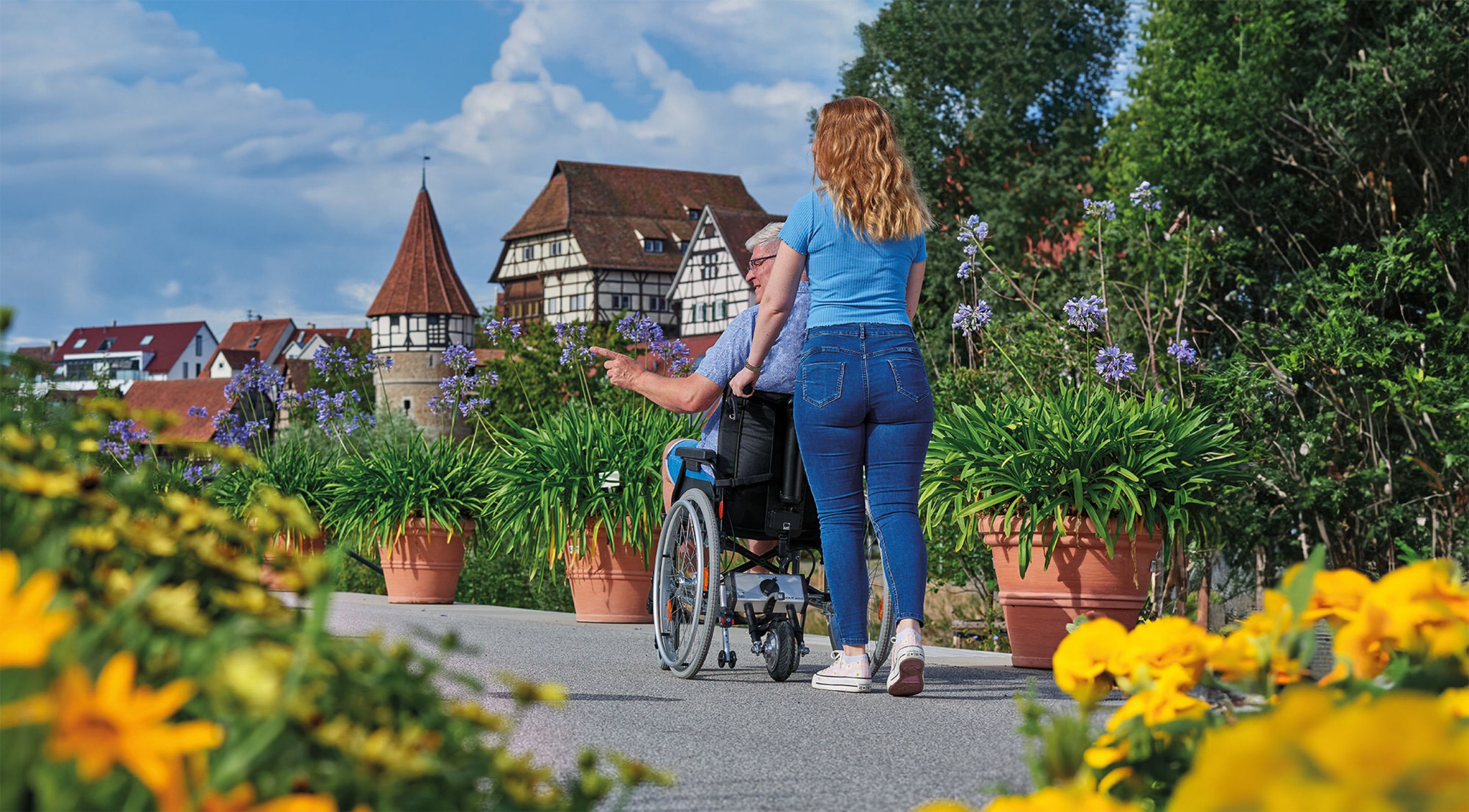 The picture shows a young woman from behind pushing an elderly man in a wheelchair up through an old town. In the foreground and background are many flowers. The wheelchair is equipped with a V-MAX mini pushing aid which supports pushing.