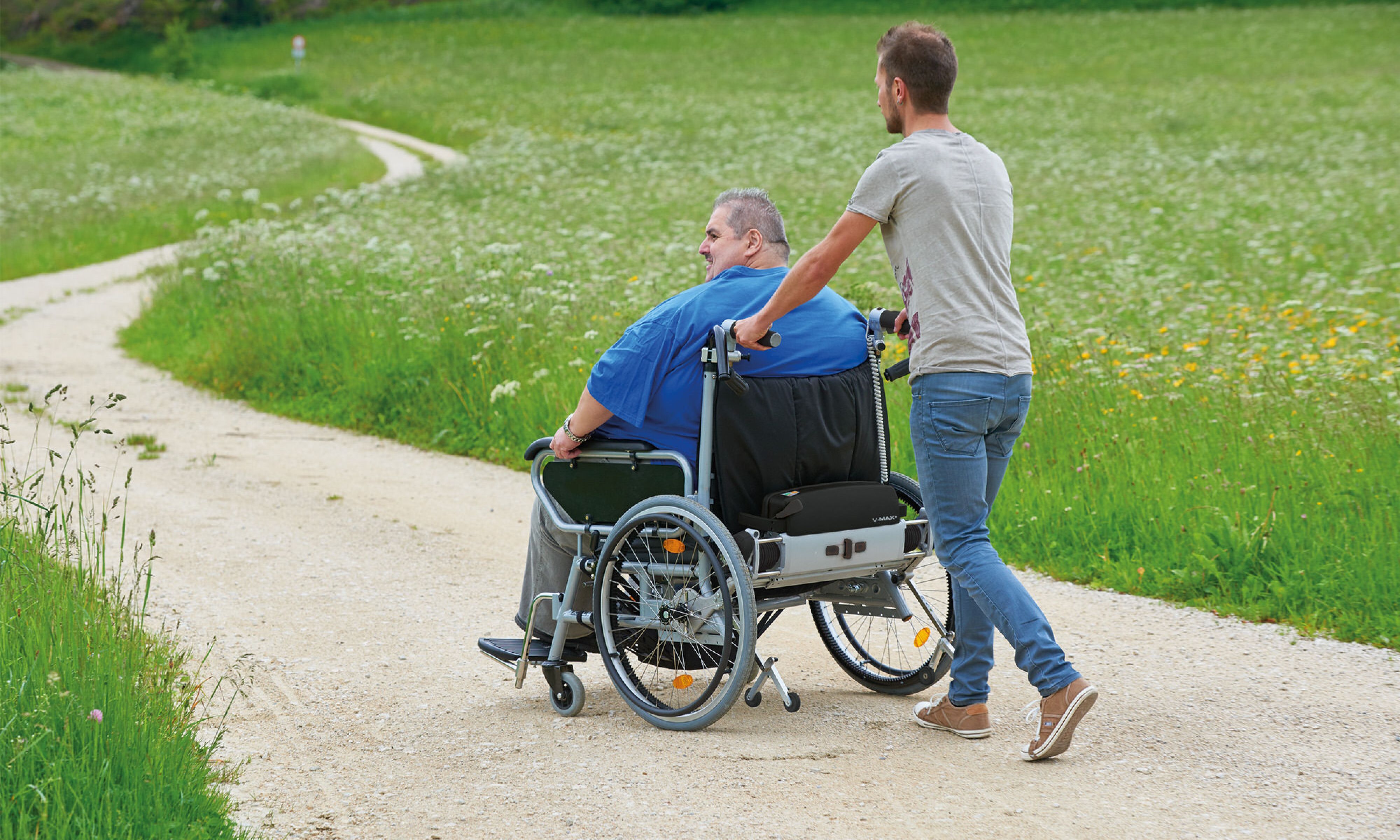 Das Bild zeigt einen sehr schlanken jungen Mann, der einen Rollstuhlfahrer mittleren Alters in einem sehr breiten Rollstuhl aufwandslos schiebt. Der Rollstuhlinsasse nutzt aufgrund seines hohen Körpergewichtes einen Rollstuhl mit Zusatzantrieb V-MAX+, der speziell für Adipositas entwickelt wurde.