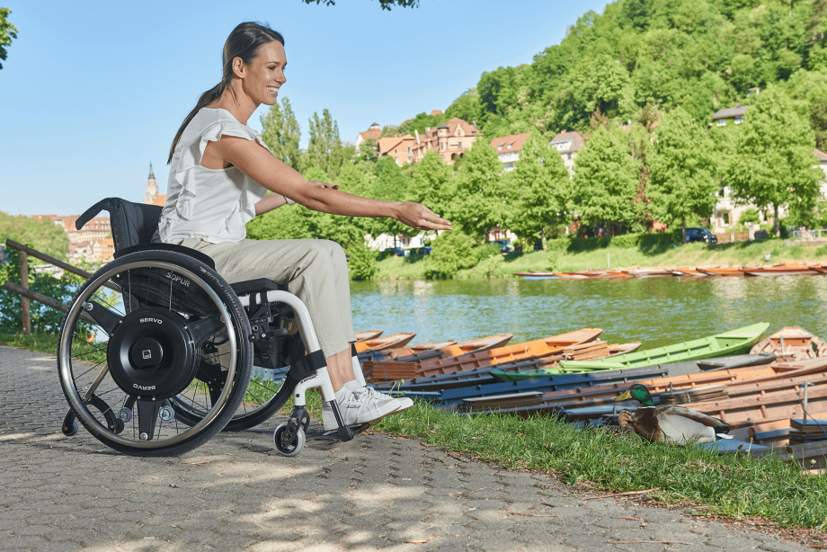 The picture shows a middle-aged woman sitting in a white wheelchair with a black wheel hub drive for boosting remaining power on the bank of a river, smiling and feeding ducks. 