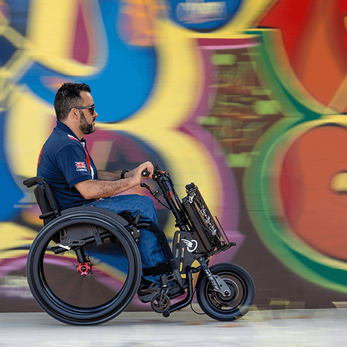 The picture shows a middle-aged man, wearing sunglasses, driving through a pedestrian zone on a summer day in his wheelchair with an auxiliary drive for self-propelled driving. The add-on drive is black and installed in the wheels as a wheel hub drive.