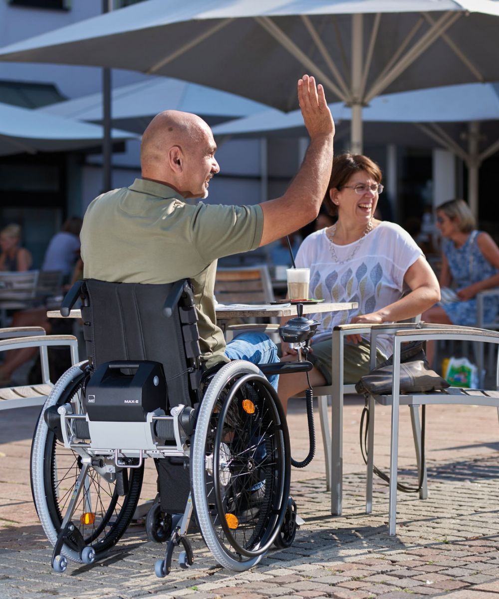 Das Bild zeigt einen Mann mittleren Alters, der mit Sonnenbrille an einem Sommertag mit seinem Rollstuhl mit Zusatzantrieb zum selbst fahren, durch eine Fußgängerzone fährt. Der Zusatzantrieb ist schwarz und in den Rädern als Radnabenantrieb verbaut.