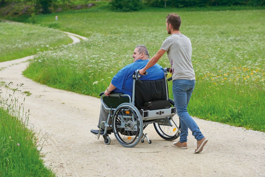 Das Bild zeigt einen sehr schlanken jungen Mann, der einen Mann mittleren Alters in einem XXL-Rollstuhl aufwandslos schiebt. Der Rollstuhlfahrer nutzt aufgrund seines hohen Körpergewichtes einen Rollstuhl mit Brems- und Schiebehilfe V-MAX+, der speziell für adipöse Patienten entwickelt wurde.