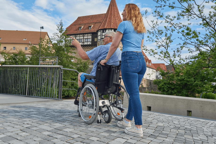 Das Bild zeigt eine junge Frau von hinten, die einen älteren Mann im Rollstuhl auf einem Weg mit Pflastersteinen schiebt. Im Hintergrund ist eine Burg zu sehen. Am Rollstuhl befindet sich hinten eine Schiebehilfe V-MAX mini, welche über das Antriebsrad beim Schieben des Rollstuhls unterstützt.
