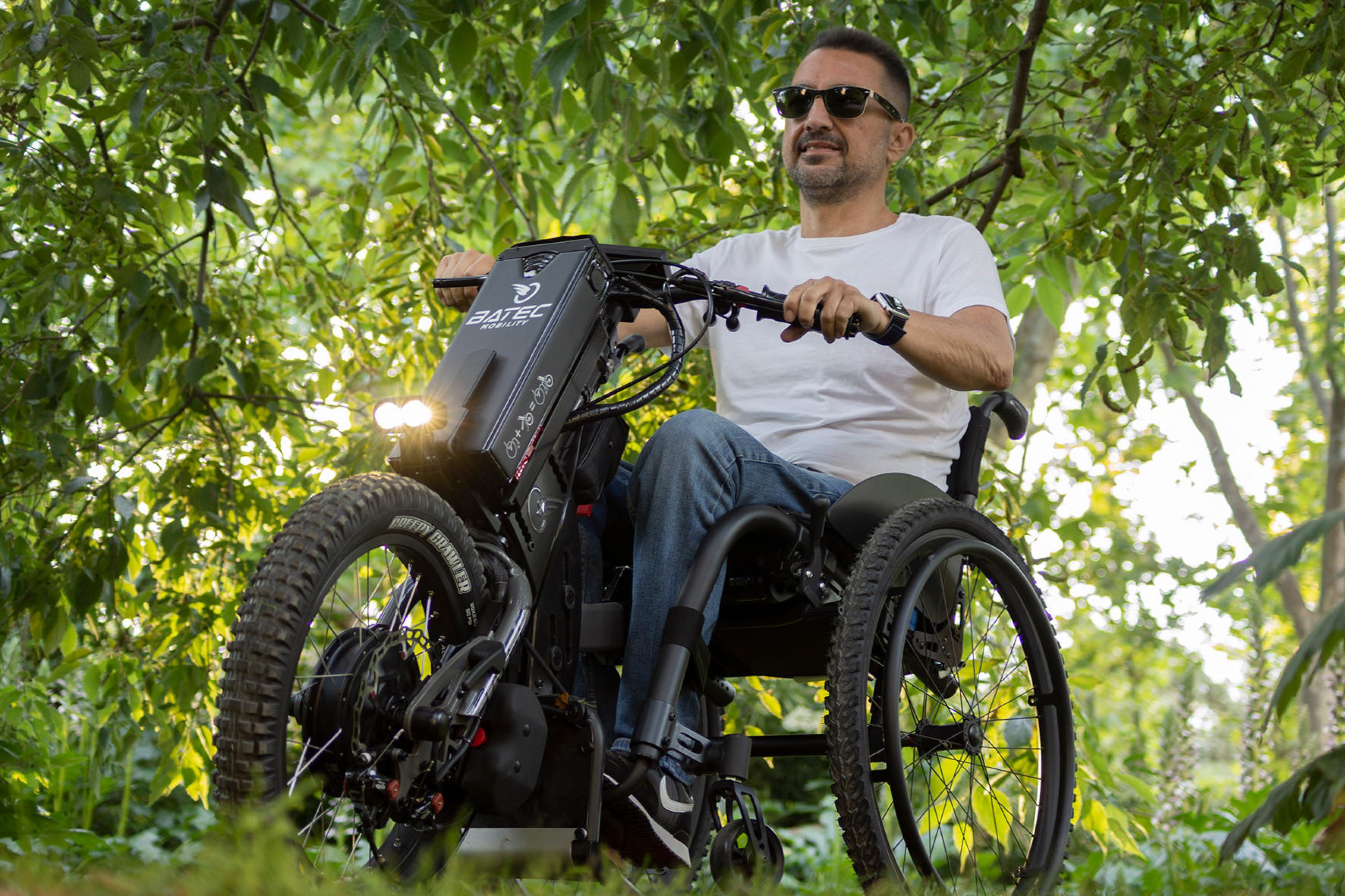 Das Bild zeigt einen Mann mittleren Alters, mit dunklen kurzen Haaren und Sonnenbrille. Er sitzt in einem grauen Rollstuhl und hat das Zuggerät BATEC Scrambler 2 vorgespannt. Er fährt an einem sonnigen Tag durch einen grünen Wald. 