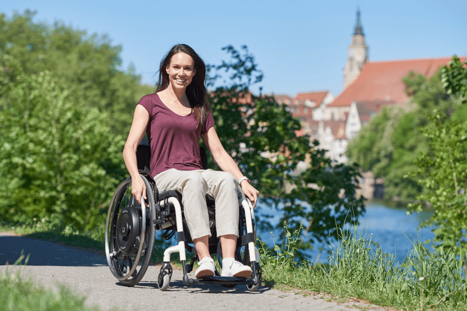 The picture shows a smiling middle-aged woman in a white wheelchair with a black wheelchair drive for residual power amplification, driving smilingly towards the camera. In the background there is a river, green trees and a city.