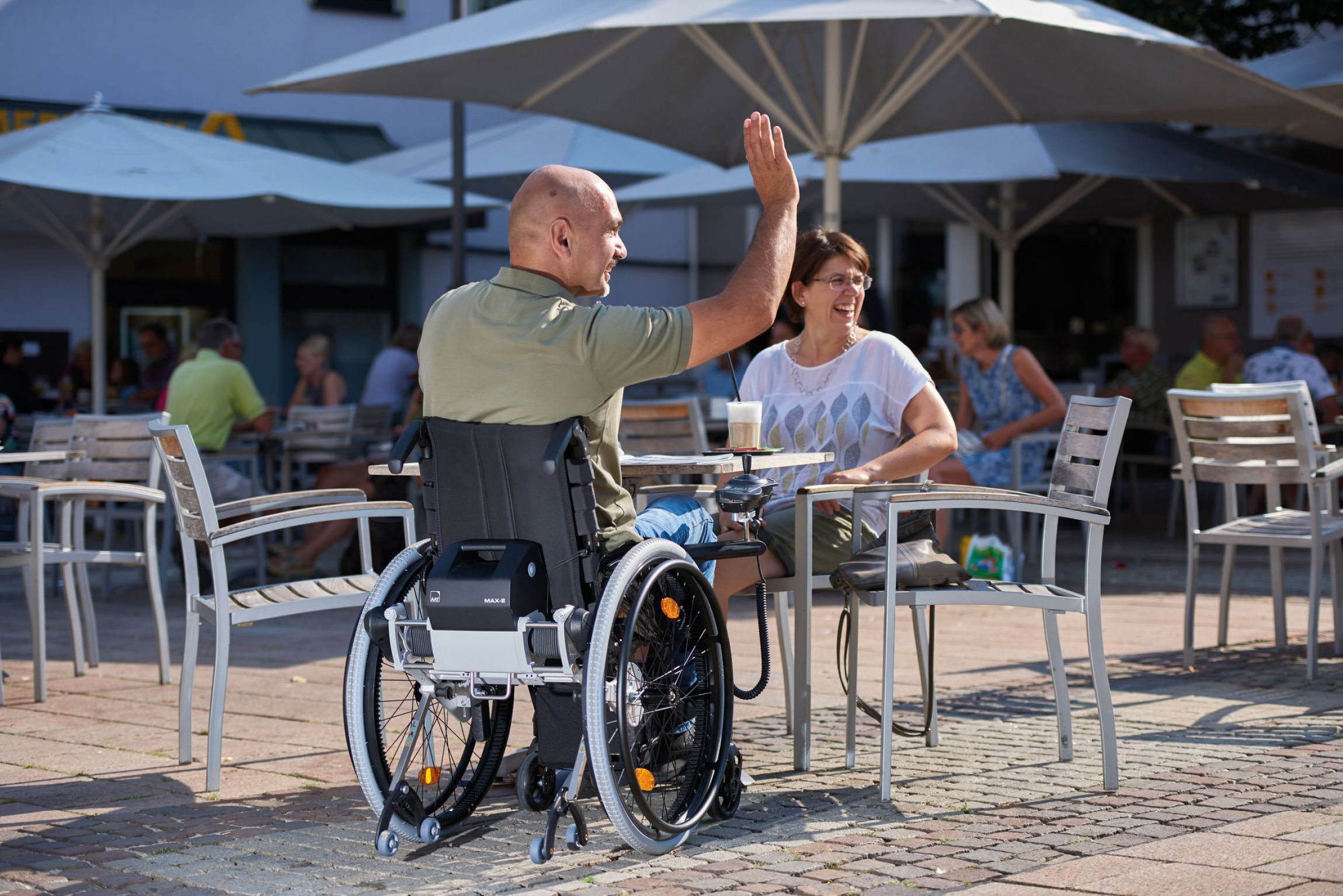 Das Bild zeigt einen Mann mittleren Alters, der im Rollstuhl sitzt und mit einer Frau in einem Cafè einen Kaffee trinkt. Beide lachen und er hält die Hand zur Begrüßung vorbeilaufender Menschen nach oben. An seinem Rollstuhl befindet sich der Zusatzantrieb MAX-E. Dieser befindet sich unterhalb der Rückenlehne seines Rollstuhls. 