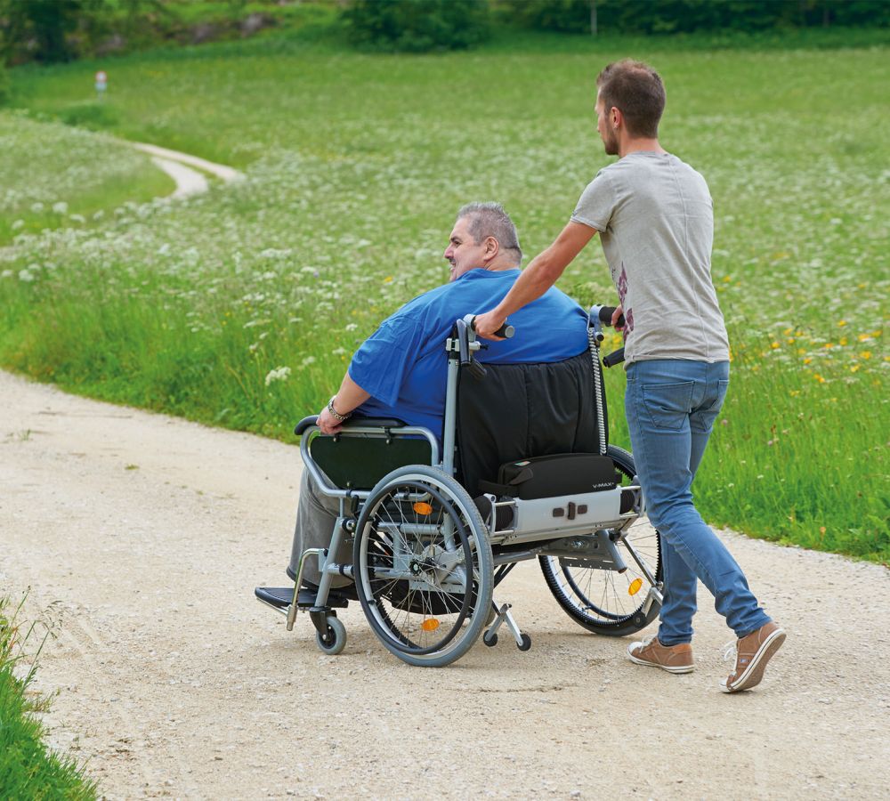 The picture shows a slim man pushing a man in a wheelchair. This man is very overweight and a V-MAX+ pushing and braking aid is used for support.