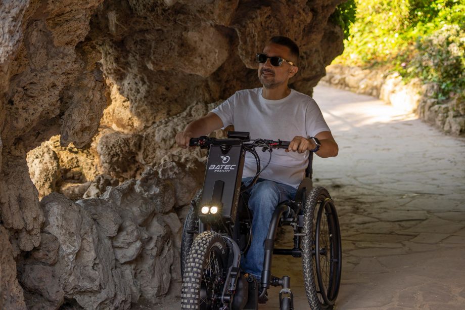 Auf dem Bild ist erneut der Mann mit Sonnenbrille zu sehen, der an einem sonnigen Tag mit seinem Rollstuhlzuggerät BATEC SCRAMBLER 2 durch einen angelegten Park fährt. Auf diesem Bild fährt er durch eine Steinhöhle, in der es dunkler ist. Die 2000-Lumen Scheinwerfer erhellen ihm die Sicht.