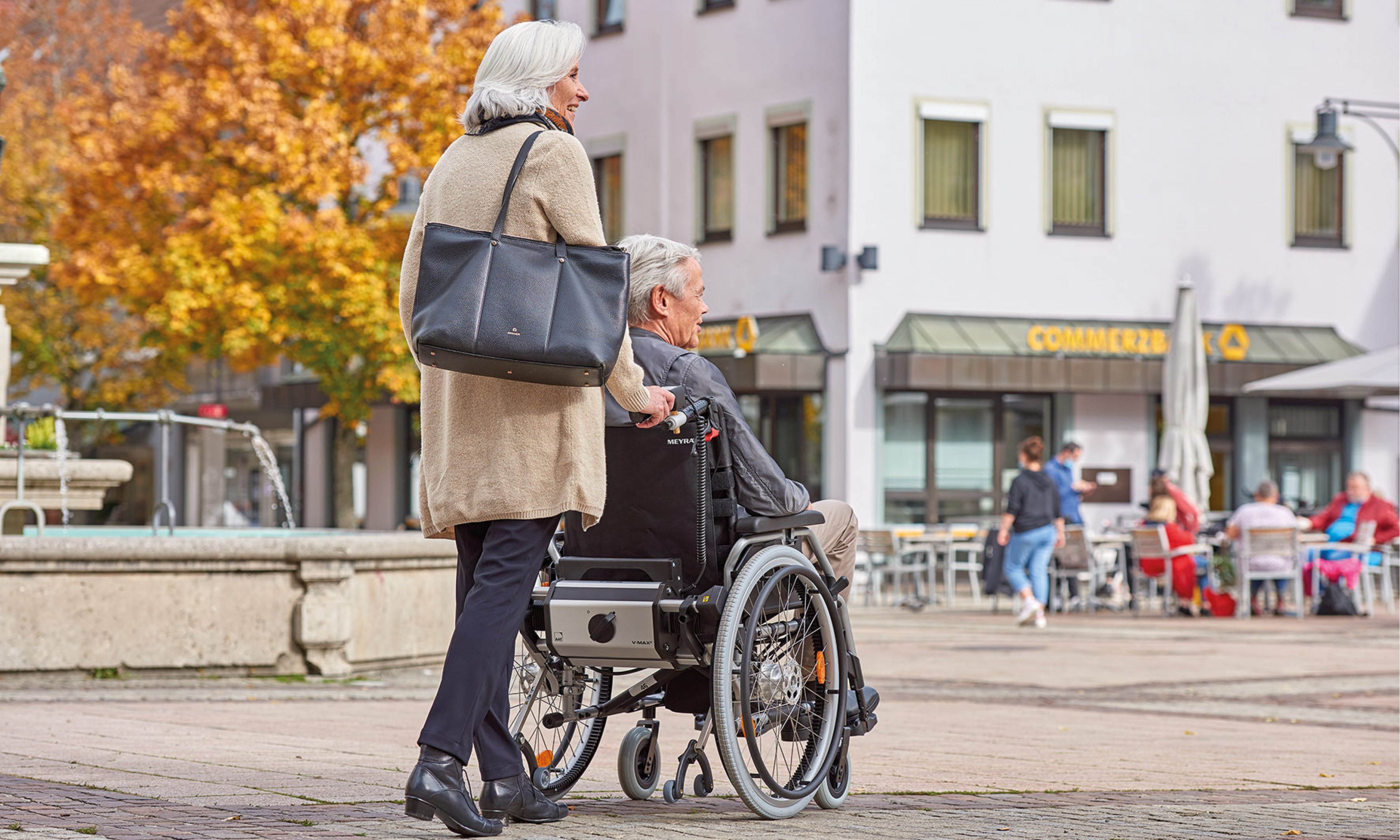 Das Bild zeigt eine ältere Frau mit grauen Haaren und großer Handtasche, wie sie einen älteren Mann mit ebenfalls grauen Haaren an einem Herbsttag in seinem manuellen Rollstuhl durch die Stadt schiebt. Am Rollstuhl befindet sich unterhalb der Rückenlehne ein AAT-Zusatzantrieb V-MAX2, welcher sie beim Schieben und Bremsen des Rollstuhls unterstützt. Sie fahren auf einem Boden, der mit unterschiedlichen Pflastersteinen ausgestattet ist.