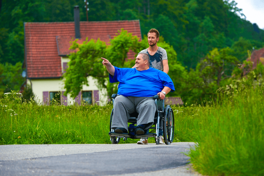 Das Bild zeigt einen sehr schlanken jungen Mann, der einen Mann mittleren Alters in einem breiten Rollstuhl ohne Kraftanstrengung schiebt. Der Rollstuhlantrieb der Firma AAT V-MAX+ wurde entwickelt, um Begleitpersonen beim Schieben und Bremsen von XXL-Rollstühlen zu helfen. 