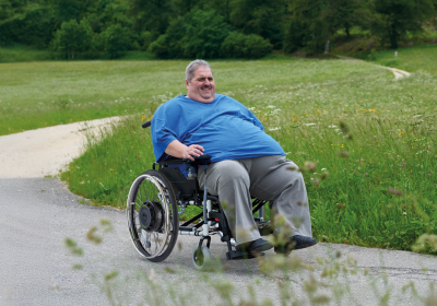 Das Bild zeigt einen Mann mittleren Alters mit einem hohen Körpergewicht. Er sitzt in einem XXL-Rollstuhl, welchen er mit einem Rollstuhlzusatzantrieb SOLO+ mit einem Bediengerät über einen Feldweg steuert. Im Hintergrund ist ein Waldrand und viel grüne Wiese sichtbar.
