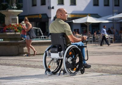 Das Bild zeigt einen Mann im Rollstuhl, der an einem sonnigen Tag einen Ausflug in die Innenstadt macht. Er nutzt einen Rollstuhl-Zusatzantrieb mit Joystick.