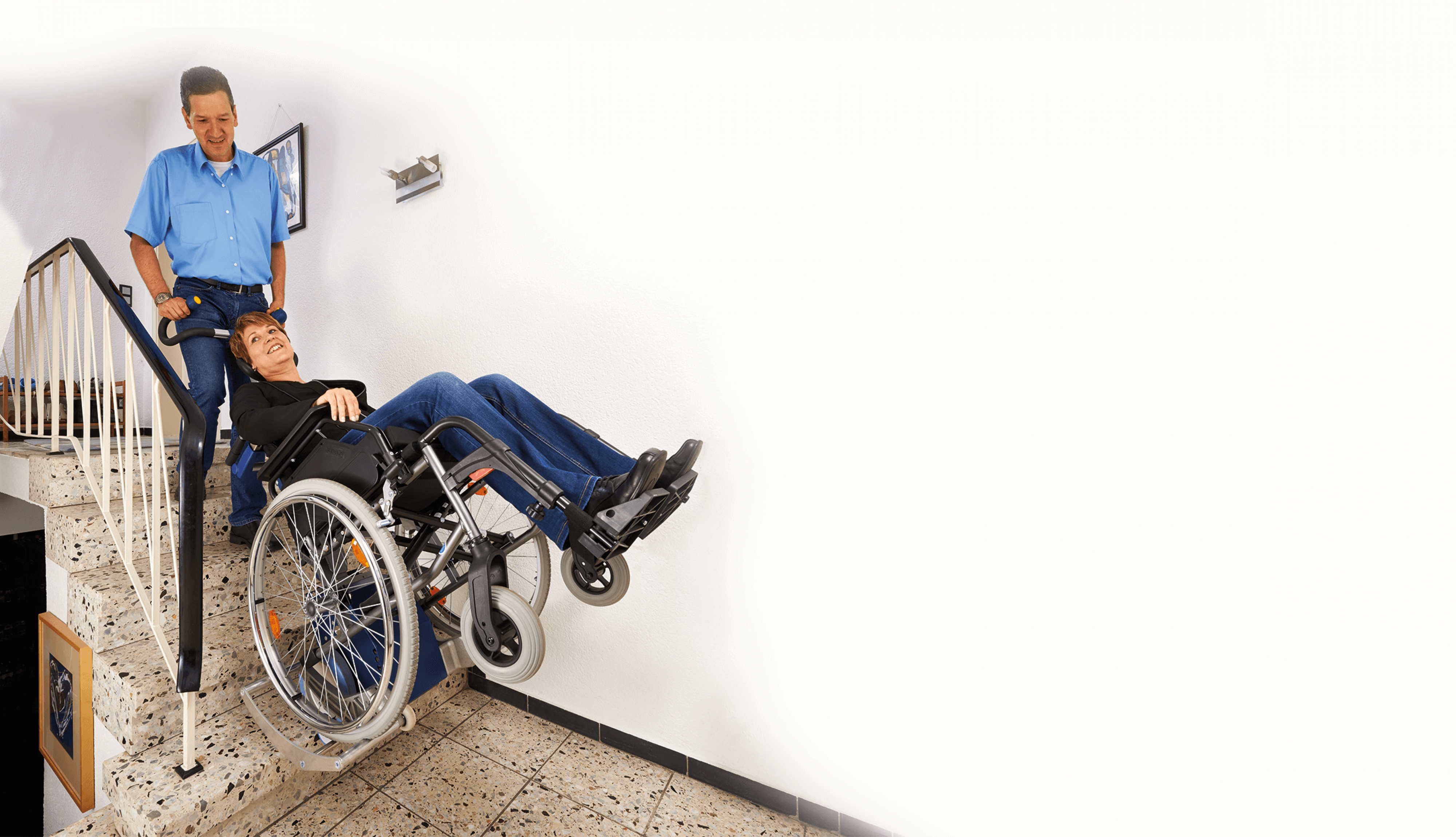 Das Bild zeigt einen Mann mittleren Alters, der eine Frau im Rollstuhl über Treppen innerhalb eines Hauses befördert. Der Rollstuhl wurde hierzu auf die Aufnahmeschuhe des Treppensteiger s-max in Kombination mit der Universalaufnahme SDM7 geschoben.