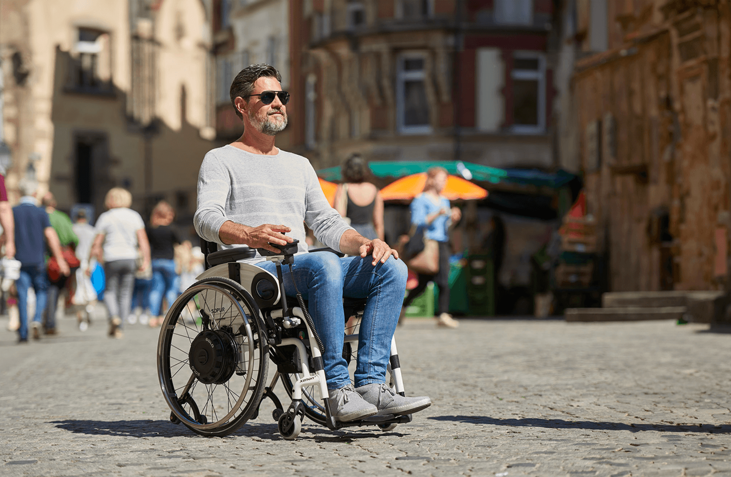 Das Bild zeigt einen Mann mittleren Alters, der mit Sonnenbrille an einem Sommertag mit seinem Rollstuhl mit Zusatzantrieb zum selbst fahren, durch eine Fußgängerzone fährt. Der Zusatzantrieb ist schwarz und in den Rädern als Radnabenantrieb verbaut.