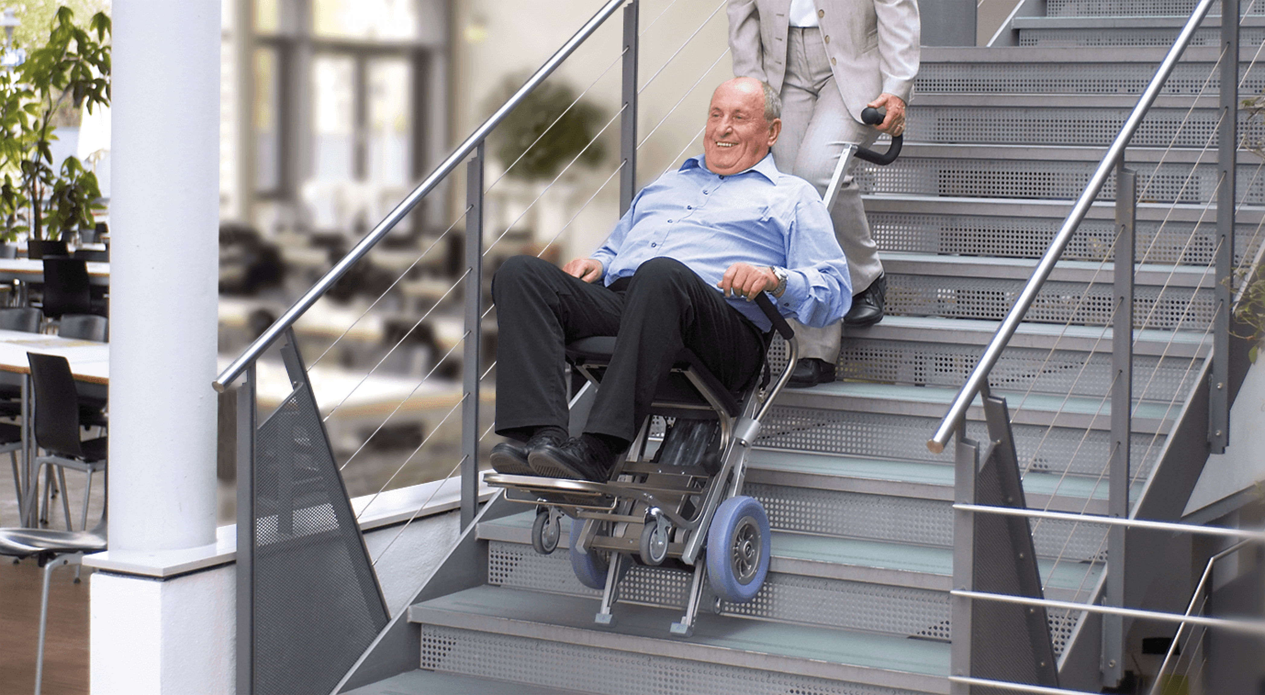Das Bild zeigt einen Mann mittleren Alters, der von einer Frau mithilfe eines Treppensteigers die Treppen hinaufbefördert wird.