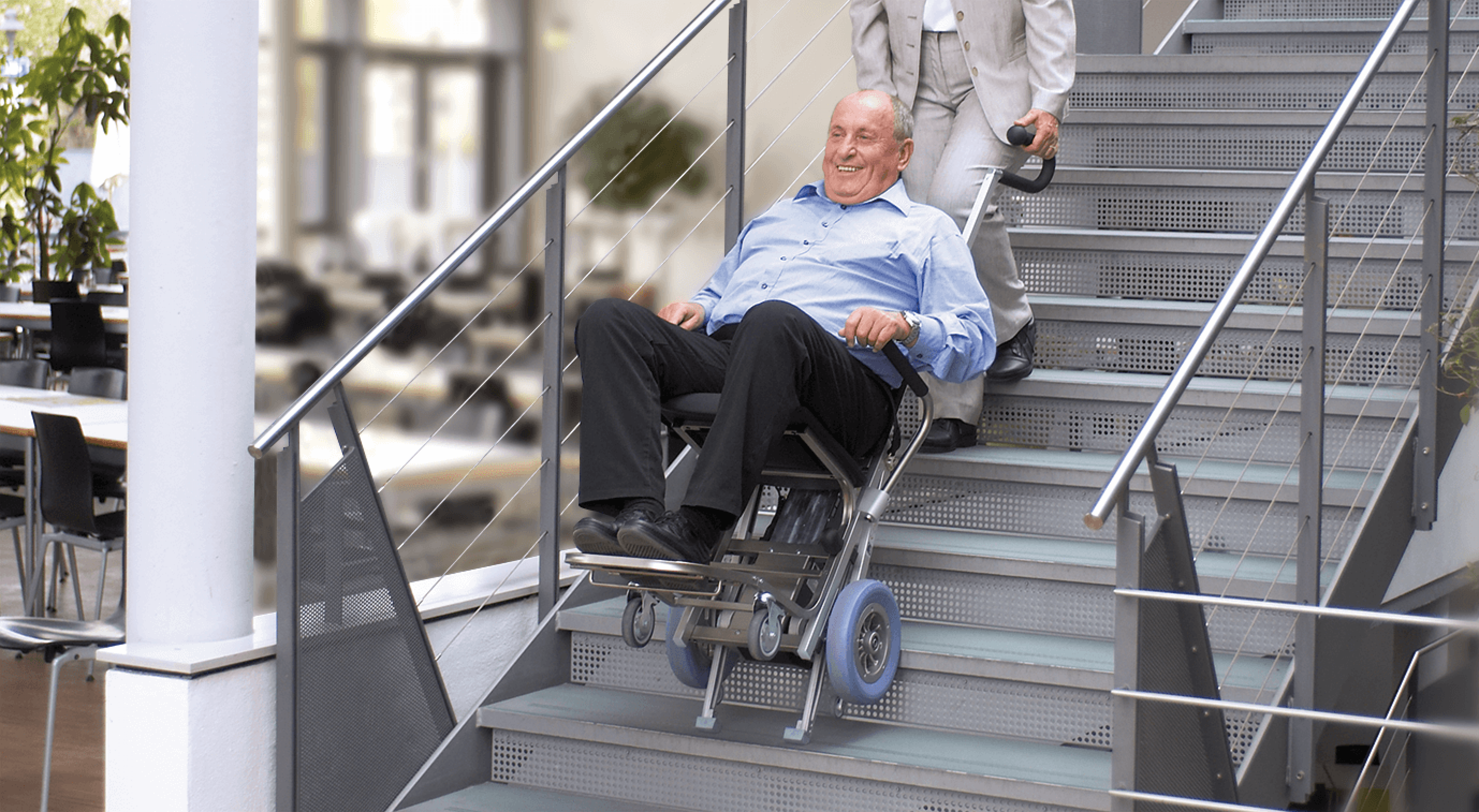 Das Bild zeigt einen Mann mittleren Alters, der von einer Frau mithilfe eines Treppensteigers die Treppen hinaufbefördert wird. 