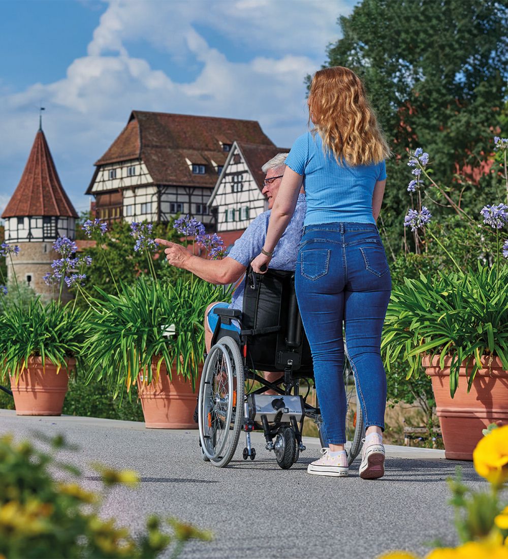 Das Bild zeigt eine junge Frau von hinten, die einen älteren Mann im Rollstuhl durch eine Altstadt hinaufschiebt. Im Vorder- und Hintergrund befinden sich viele Blumen. Am Rollstuhl befindet sich hinten eine Schiebehilfe V-MAX mini, welche über das Antriebsrad beim Schieben unterstützt.