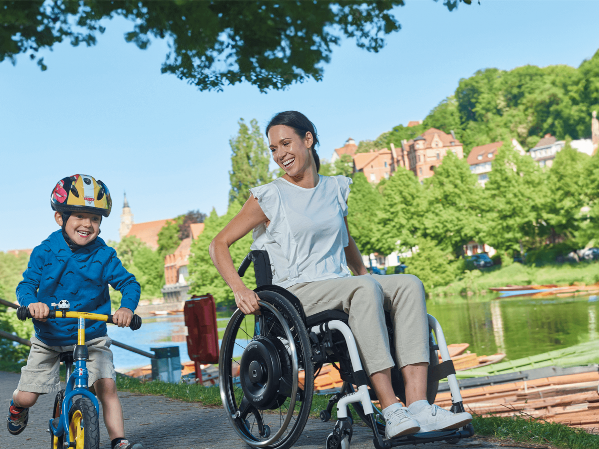 The picture shows a middle-aged man, wearing sunglasses, driving through a pedestrian zone on a summer day in his wheelchair with an auxiliary drive for self-propelled driving. The add-on drive is black and installed in the wheels as a wheel hub drive.