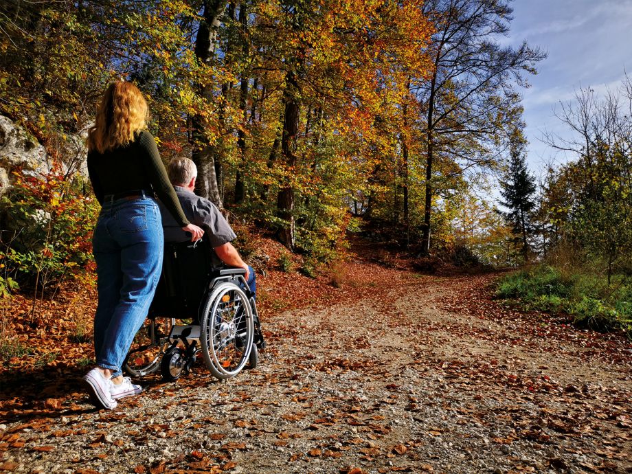 Das Bild zeigt eine junge Frau von hinten, die einen älteren Mann im Rollstuhl einen Schotterweg im Wald hinaufschiebt. Die Bäume sind bunt und es liegt Laub auf dem Weg. Am manuellen Rollstuhl befindet sich hinten eine AAT Schiebehilfe V-MAX mini, welche über das Antriebsrad der Begleitperson beim Anschieben des Rollstuhls hilft.