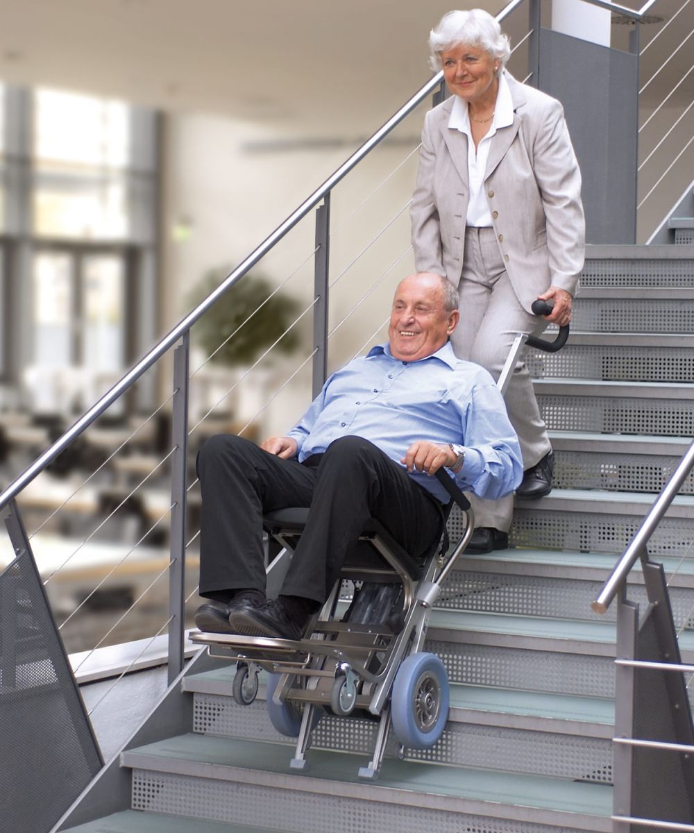 Das Bild zeigt einen Mann mittleren Alters, der von einer Frau mithilfe eines Treppensteigers die Treppen hinaufbefördert wird.