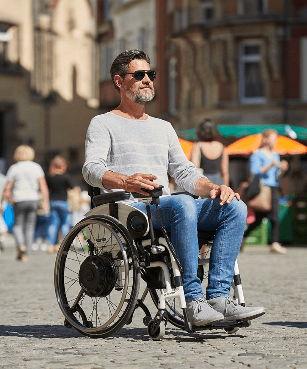 Das Bild zeigt einen Mann mittleren Alters, der mit Sonnenbrille an einem Sommertag mit seinem Rollstuhl mit Zusatzantrieb zum selbst fahren, durch eine Fußgängerzone fährt. Der Zusatzantrieb ist schwarz und in den Rädern als Radnabenantrieb verbaut.