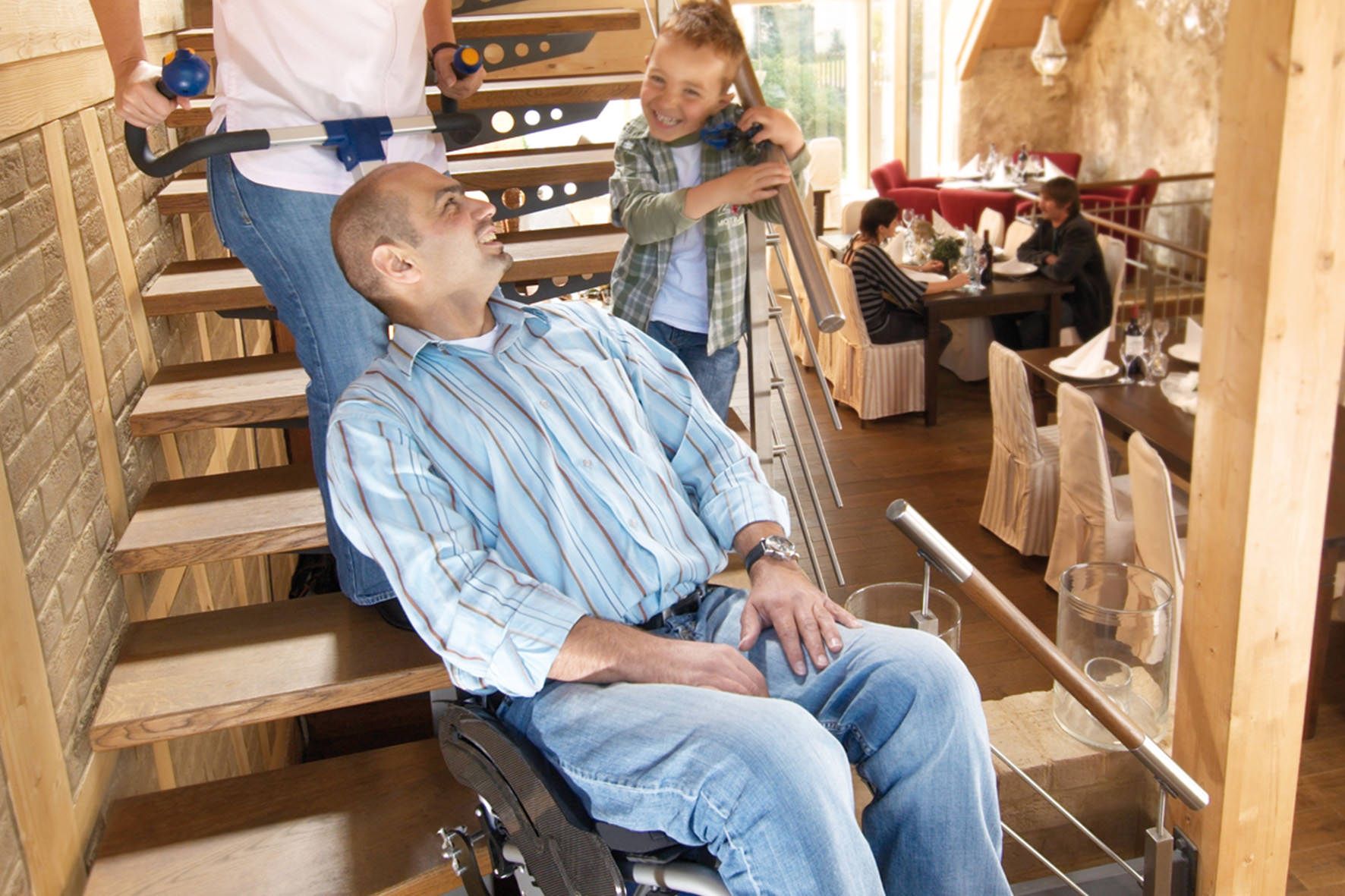 Das Bild zeigt einen Familienvater, der in einem Treppensteiger mit Rollstuhl von einer Frau über eine Treppe transportiert wird. Sein Sohn steht neben ihm auf der Treppe und lächelt ihn an.