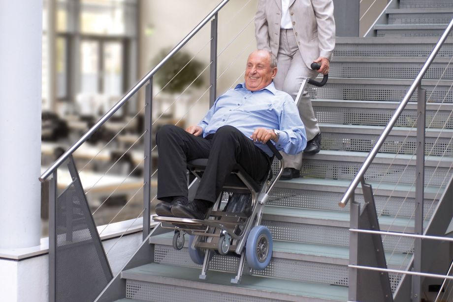 Das Bild zeigt einen Mann mittleren Alters, der von einer Frau mithilfe eines Treppensteigers die Treppen hinaufbefördert wird.