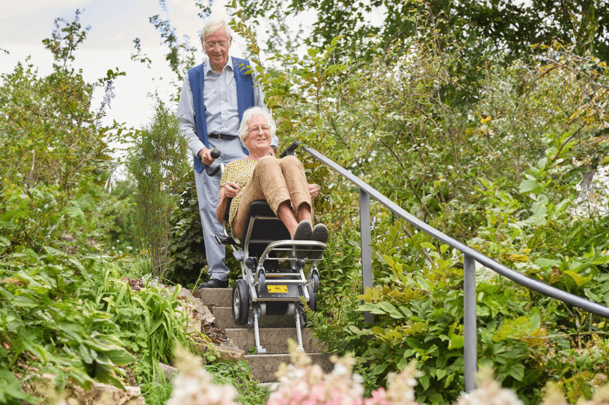 Das Bild zeigt einen älteren Herren, der in einem Treppensteiger mit integrierter Sitzeinheit von einer Frau über eine Treppe transportiert wird.