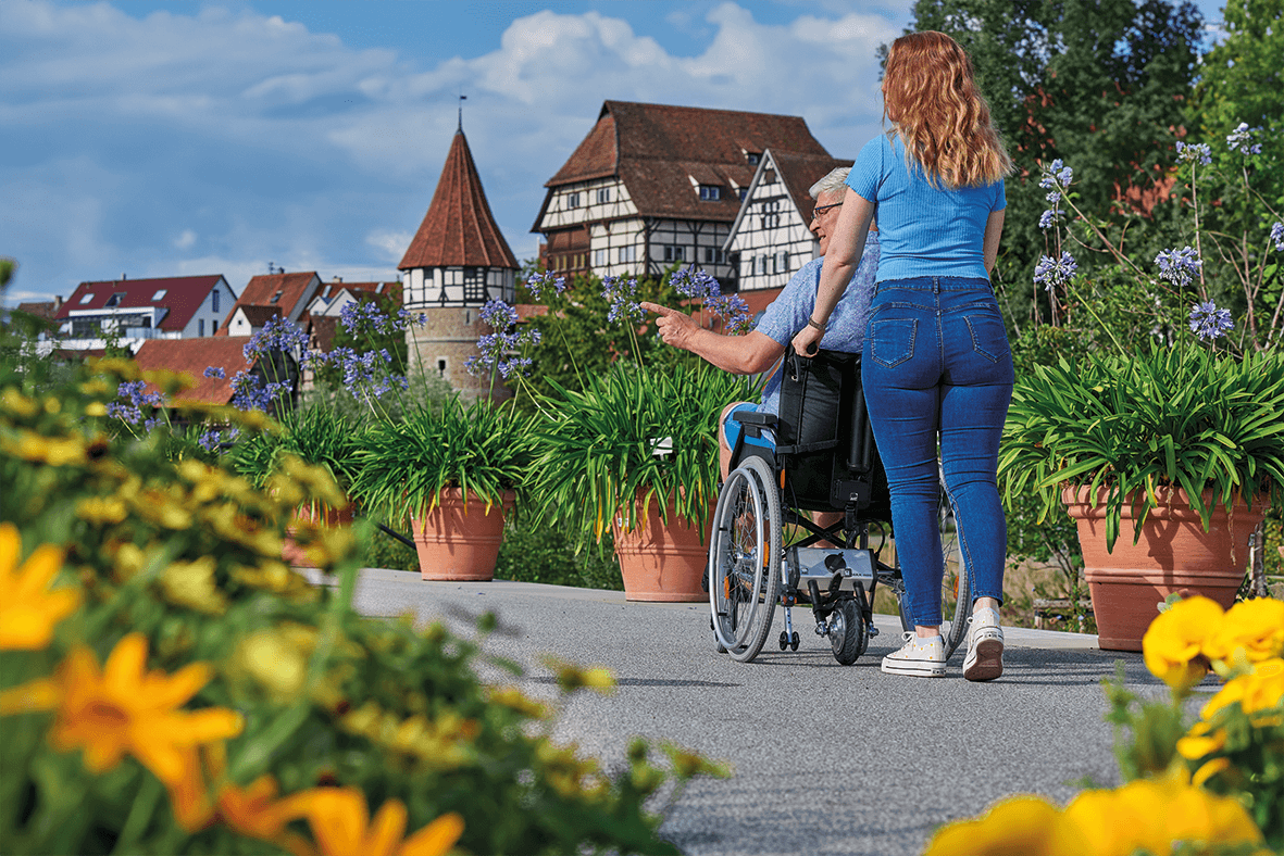 Das Bild zeigt eine junge Frau, die mit einen Mann einen Ausflug in die Natur macht. Sie nutzt zur Entlastung eine Schiebehilfe für den Rollstuhl.